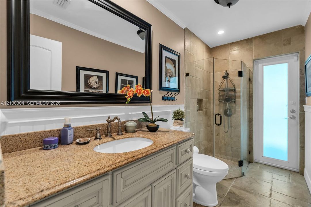Bathroom vanity featuring a stone countertop with an undermount oval sink, wooden cabinetry, large wall-mounted mirror, glass-enclosed shower with stone wall tile, and a door with a frosted glass pane