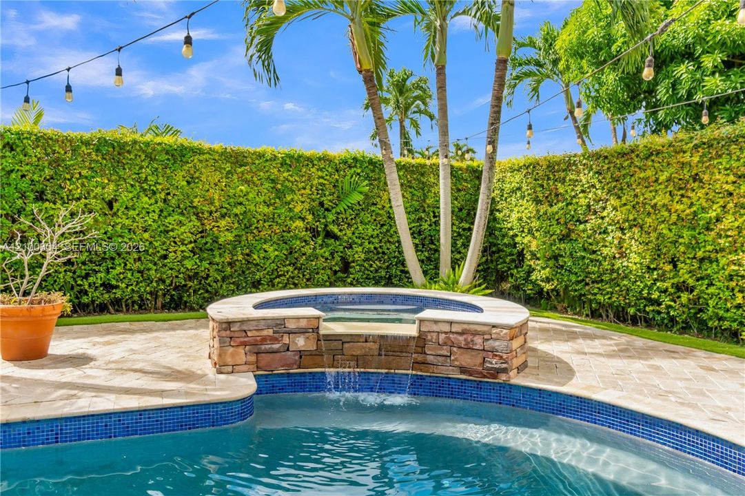 In-ground pool featuring a raised stone-veneer spa with a waterfall spillway, stone paver decking, and a perimeter privacy hedge