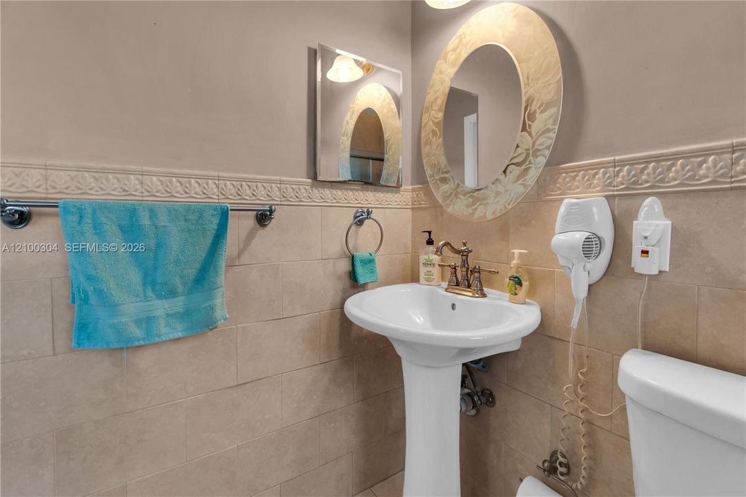 White pedestal sink with a metallic dual-handle faucet, oval wall mirror with an etched border, and neutral-toned wall tiling featuring a decorative relief trim
