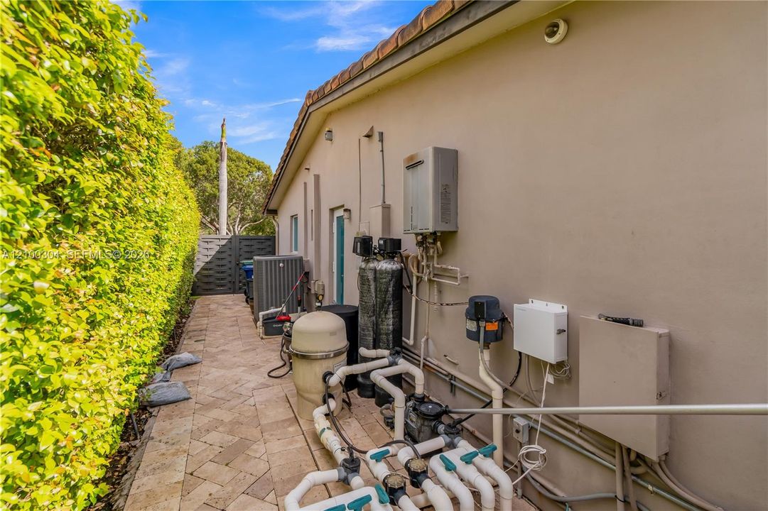 Exterior side yard featuring patterned paver flooring, a tankless water heater, water filtration system, and a swimming pool filtration unit