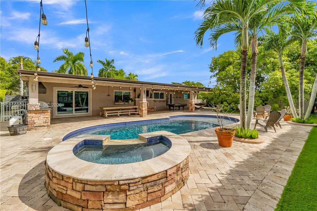 Outdoor living area featuring a covered lanai, stone-clad spa, swimming pool, herringbone-patterned stone pavers, and mature palm trees