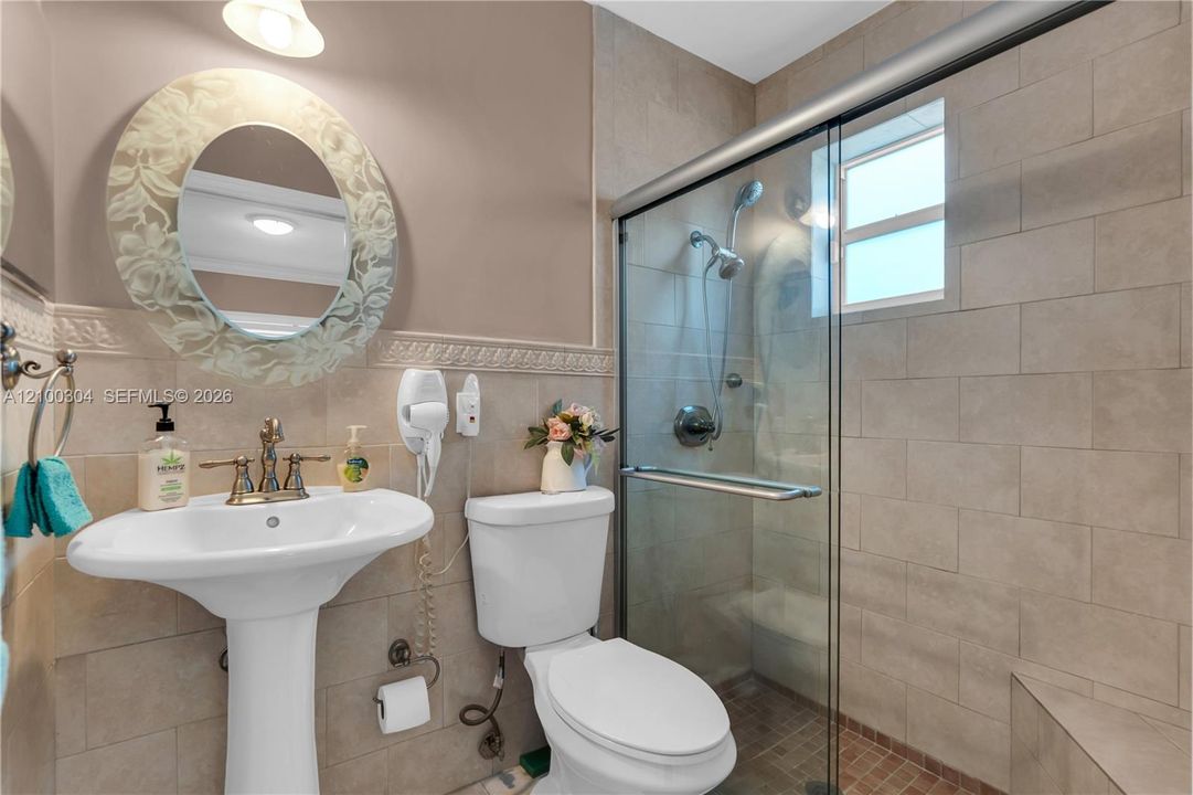 Pedestal sink with widespread faucet, wall-mounted oval mirror, ceramic wall tile, and glass-enclosed shower with adjustable showerhead