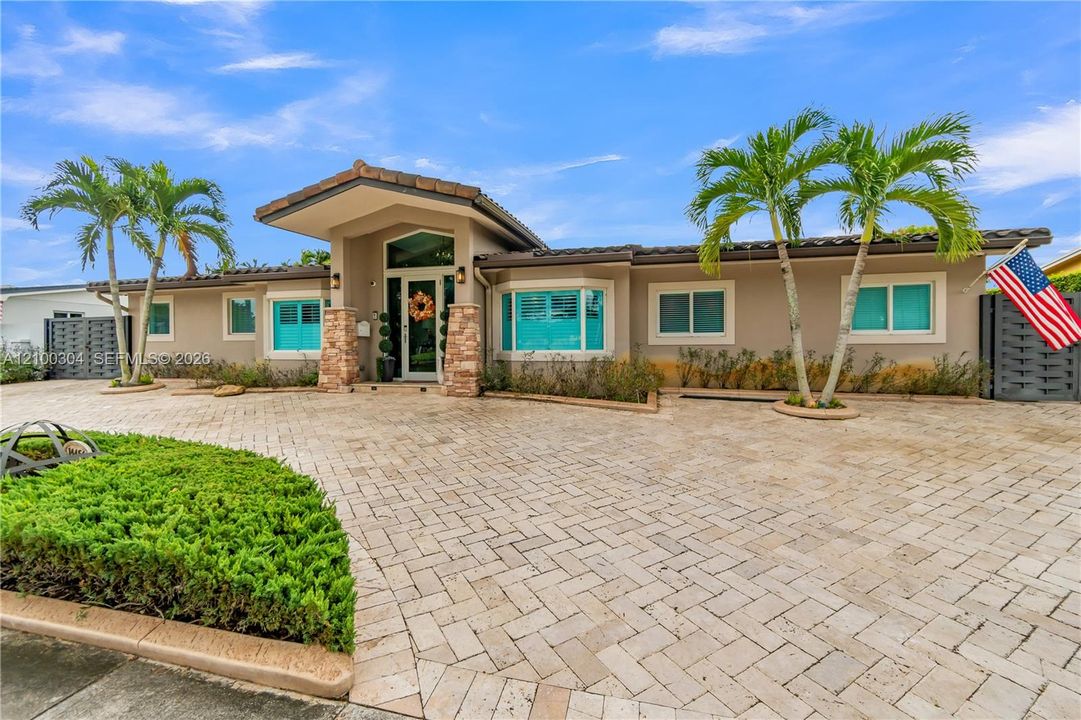 Single-story residence featuring a stone-clad portico, herringbone-patterned paved driveway, manicured landscaping, and palm trees