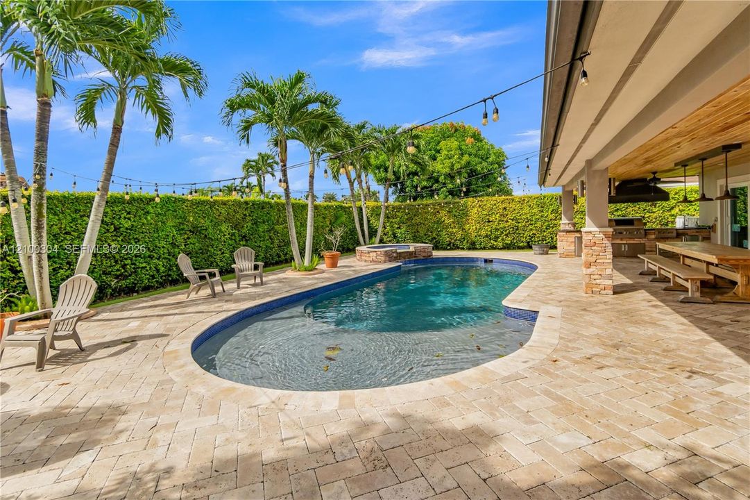 Outdoor living area featuring a swimming pool with integrated spa, herringbone-pattern stone patio, covered lanai with wood-paneled ceiling, stone-clad structural columns, and built-in outdoor kitchen