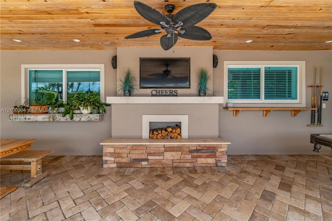 Covered patio featuring natural wood-plank ceiling, herringbone-patterned stone paver flooring, built-in stone fireplace with hearth, and two wood-shelf window ledges