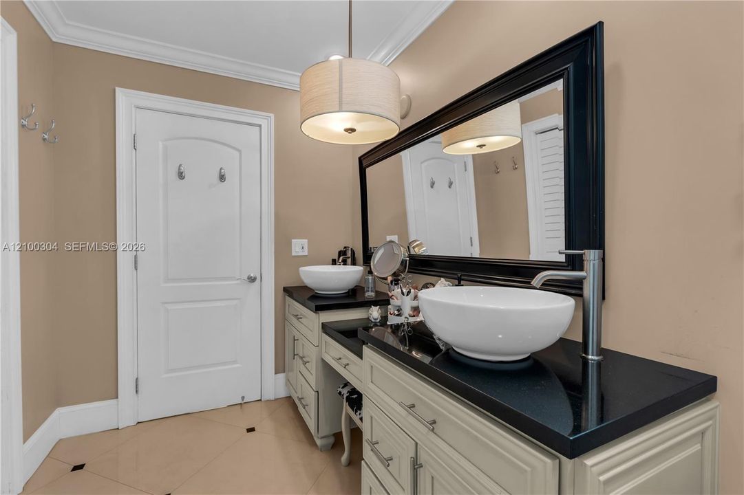 Dual vessel sinks with polished chrome faucets, black stone countertop, cream-colored cabinetry, and large framed wall mirror