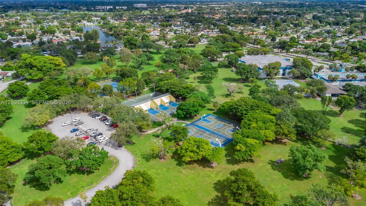 Aerial perspective of park grounds featuring a paved parking area, basketball courts, tennis courts, and mature landscaping