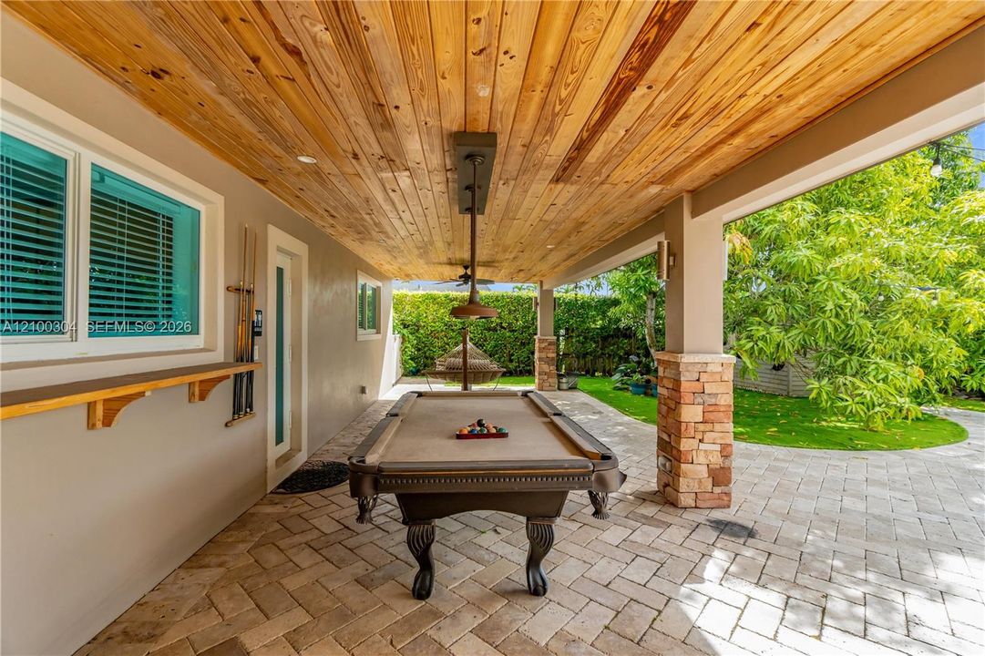 Covered patio featuring a wood-plank ceiling, natural stone column accents, herringbone-patterned paver flooring, and an attached exterior wall-mounted shelf