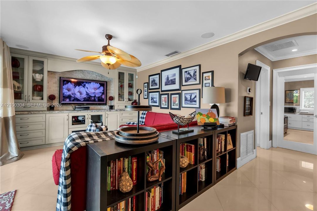 Custom built-in cabinetry featuring recessed-panel doors, neutral stone countertop, stone tile backsplash, and integrated light fixtures, complemented by polished tile flooring and a ceiling-mounted fan with palm-style blades