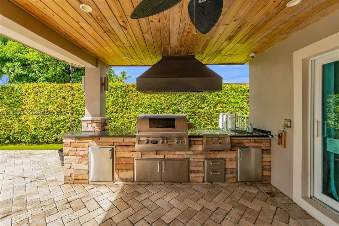 Covered outdoor kitchen featuring a stone-clad base, stainless steel built-in grill and storage, dark stone countertop, and wood-paneled ceiling, all set on a herringbone-patterned stone paver patio