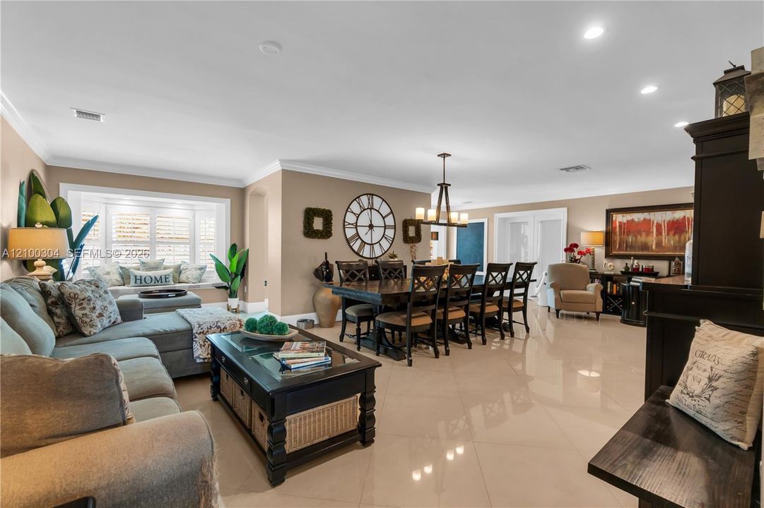 Open-concept area featuring polished tile flooring, crown molding, a built-in window seat with plantation shutters, recessed lighting, and a multi-light chandelier