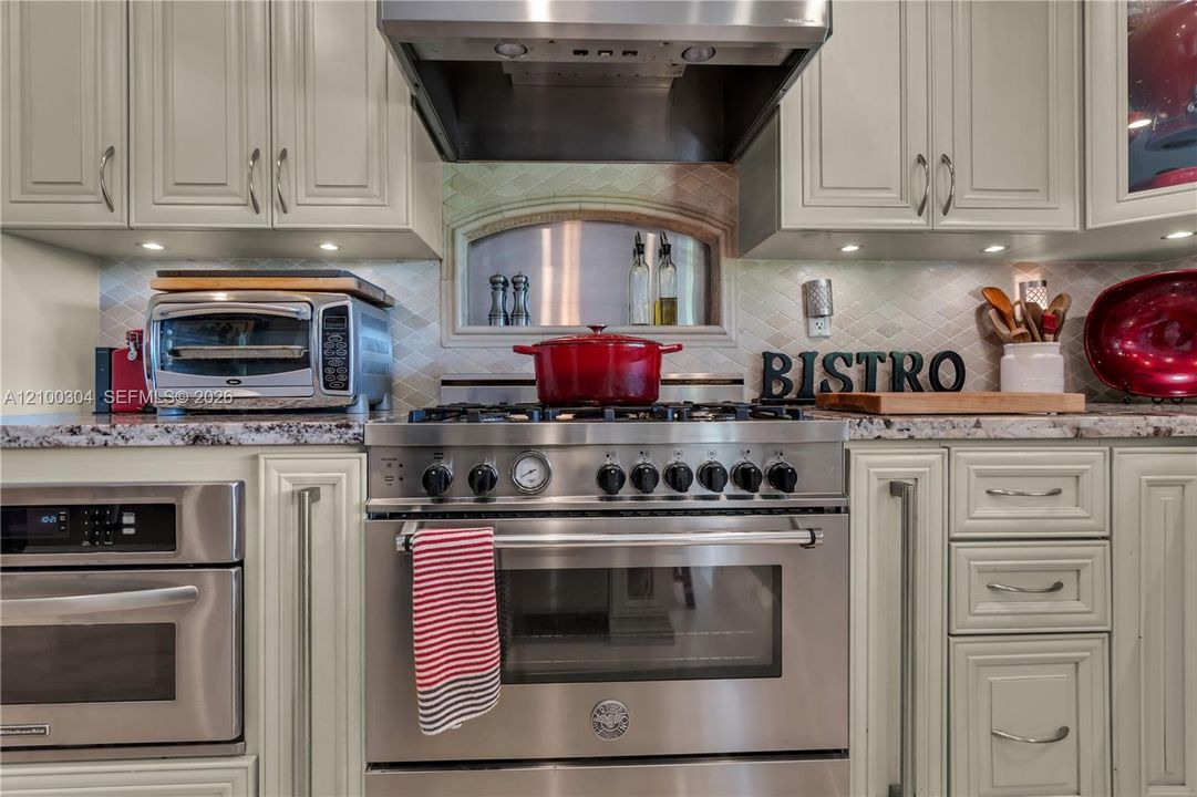 Kitchen featuring professional-grade stainless steel gas range, built-in wall oven, stone countertops, custom cabinetry with under-cabinet lighting, and a decorative arched backsplash niche