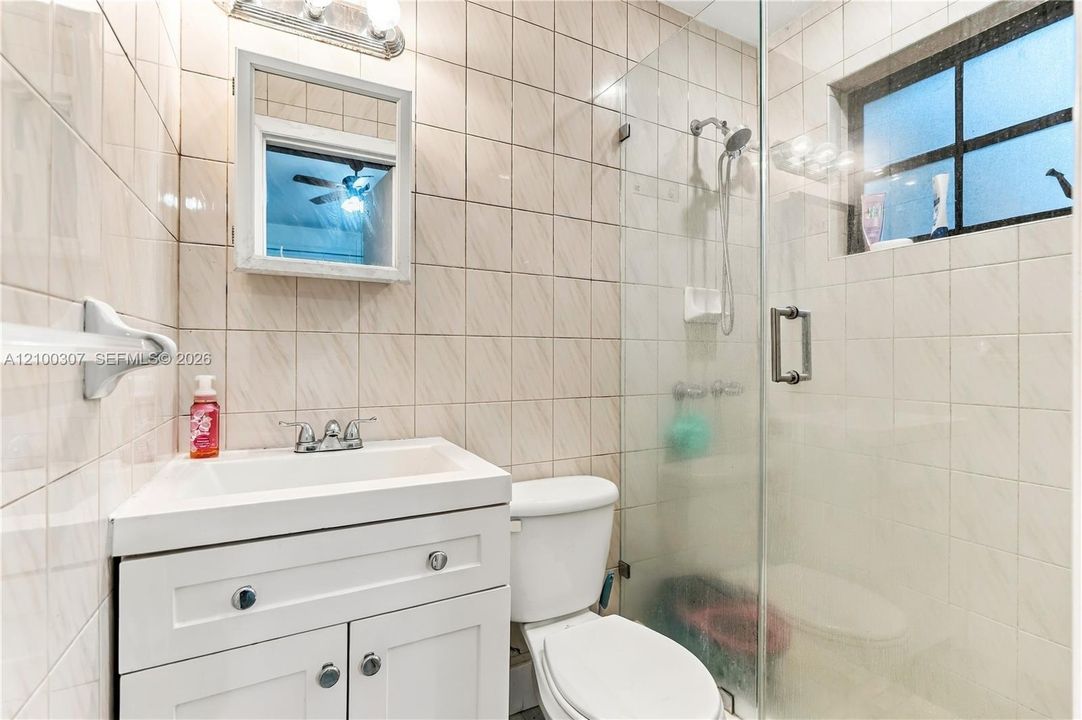Full-tiled bathroom featuring a white vanity cabinet with a drop-in sink, glass-enclosed shower, wall-mounted medicine cabinet, and grid-pattern window