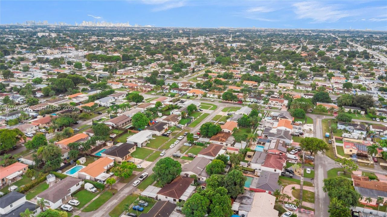Aerial perspective featuring a grid-patterned neighborhood with mature trees, paved roadways, and urban skyline visibility on the horizon