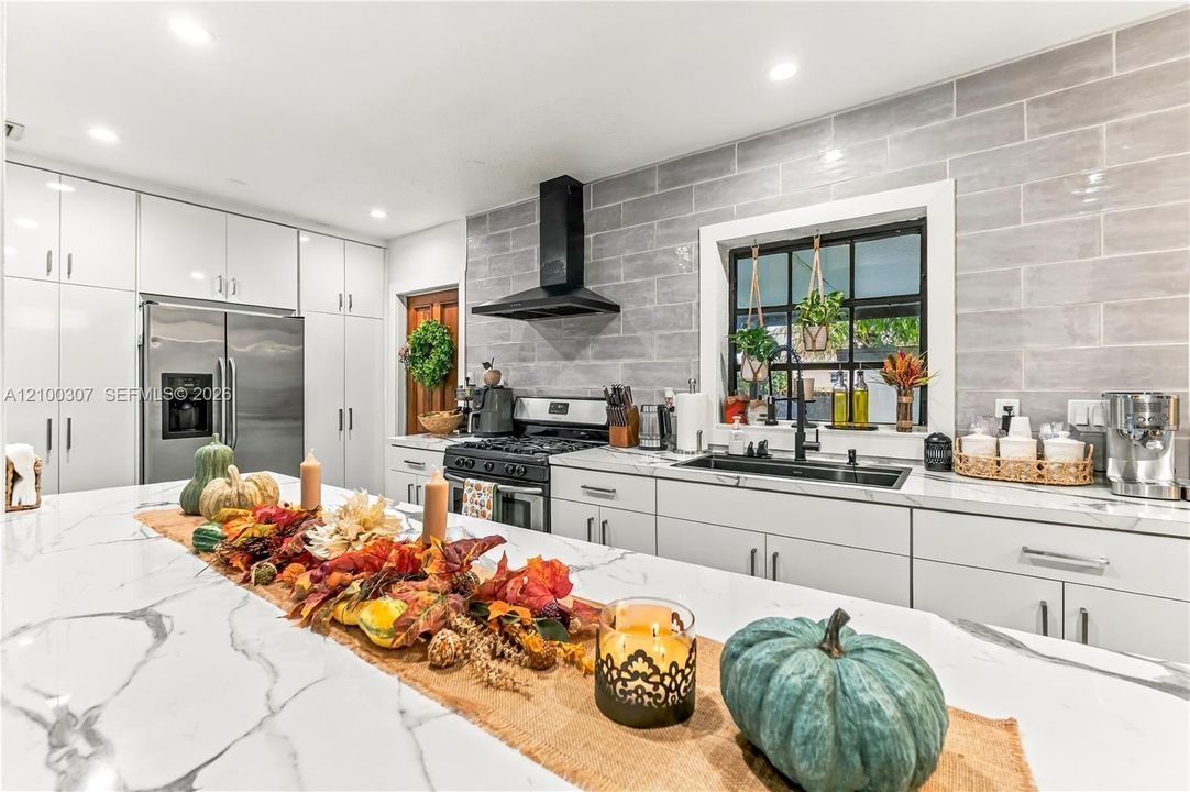 Kitchen featuring white flat-panel cabinetry, a stone-patterned waterfall edge island, subway tile backsplash, matte black range hood, and black-framed window