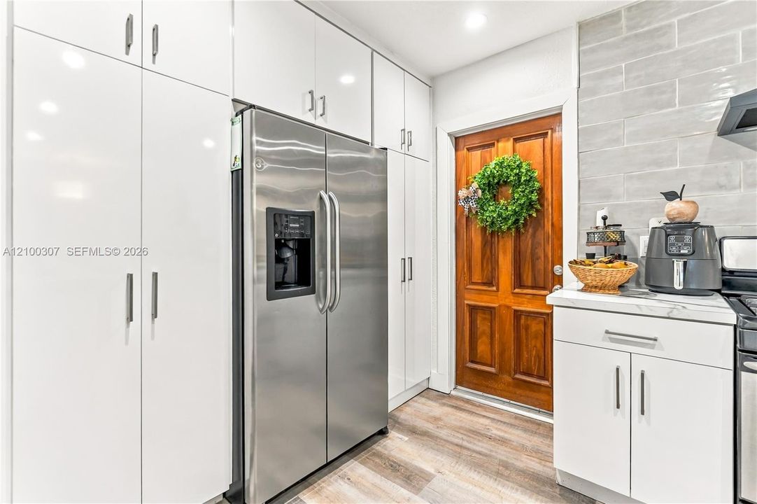 Kitchen featuring white high-gloss cabinetry, stainless steel side-by-side refrigerator, wood-finish flooring, grey subway-style tile backsplash, and a six-panel wood door