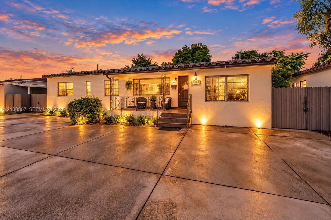 Spanish-style exterior featuring smooth stucco siding, a barrel-tiled roof, an elevated concrete porch with decorative metal railing, a wide concrete driveway, and solid wood-finish fencing