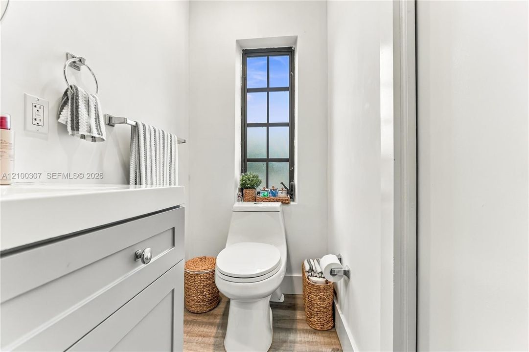 Modern vanity with a light gray finish, wood-finish flooring, white vitreous china toilet, and a multi-pane grid window
