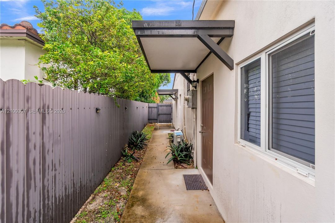 Side exterior featuring a concrete walkway, corrugated metal fencing, metal door awnings, and white stucco exterior finish