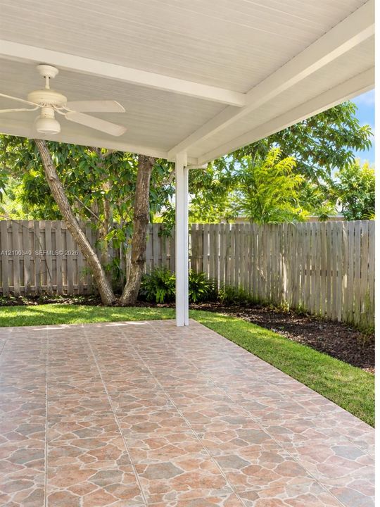 Covered patio featuring stone-patterned tile flooring, a white ceiling fan, wood fencing