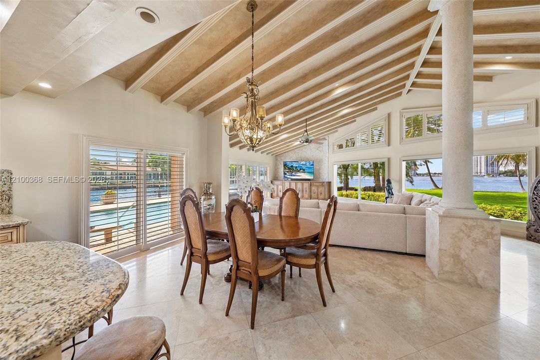 Open-concept living space featuring vaulted ceilings with exposed wood beams, natural stone flooring, a decorative stone column, and expansive floor-to-ceiling windows with waterfront exposure
