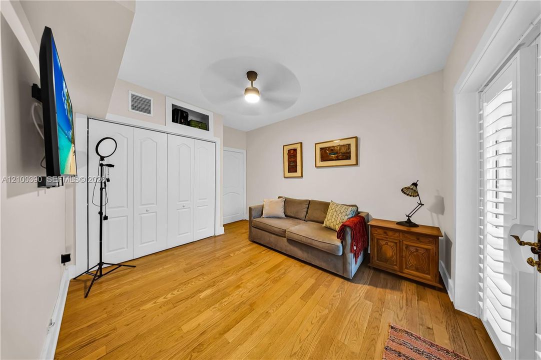 Room featuring wood-finish flooring, white bi-fold closet doors with built-in overhead storage, a ceiling fan with integrated lighting, and white plantation shutters on a glass-paned door