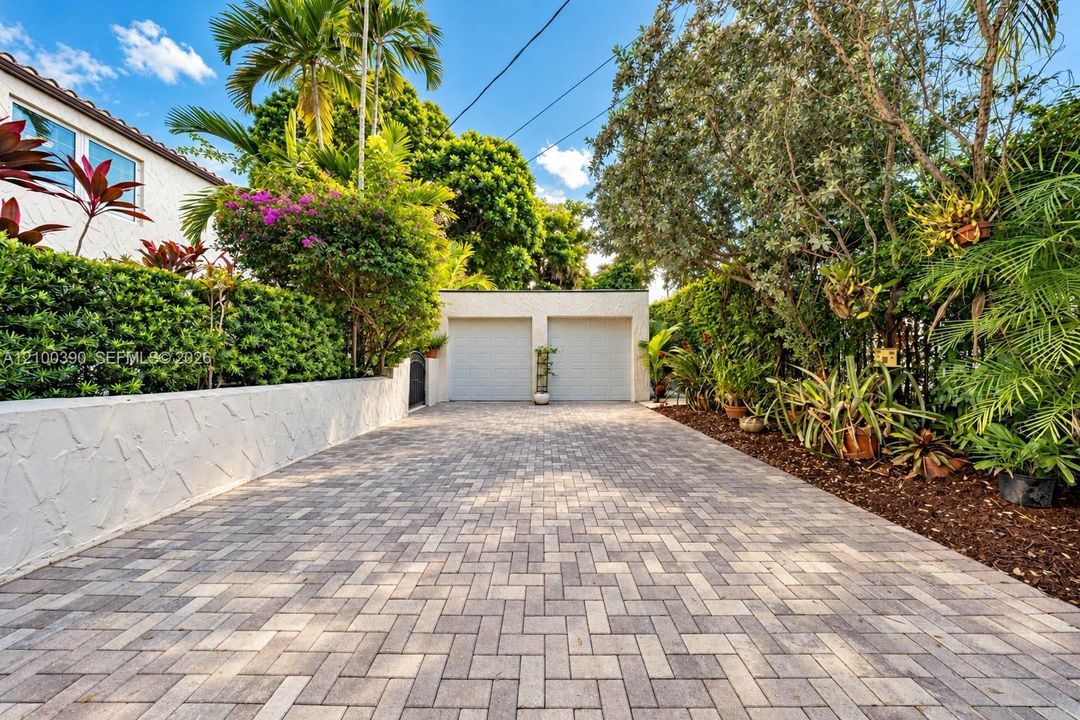 Paver driveway featuring a detached two-car garage, white stucco privacy wall, and mature landscaping