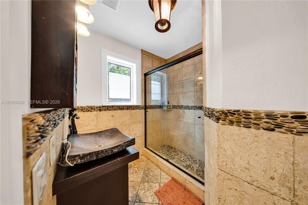 Stone vessel sink with dark finish, natural stone wall tile with pebble accent border, glass-enclosed shower with pebble flooring, and metal pendant lighting