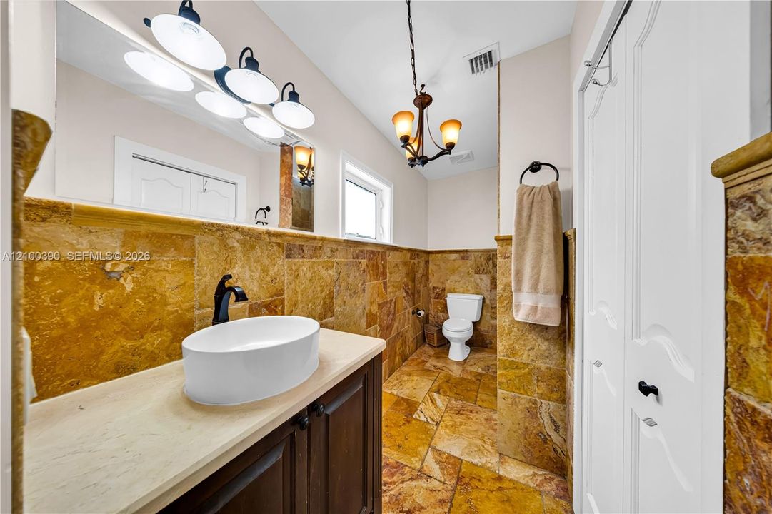 Stone-tiled bathroom featuring a vessel sink with a dark-finish faucet, stone countertop, custom cabinetry, and decorative overhead lighting