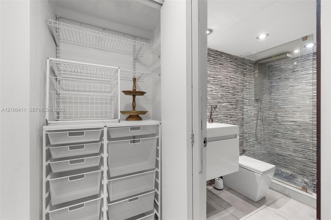 Walk-in closet featuring wire shelving and a modular storage system, paired with a bathroom containing a wall-mounted vanity, square-profile toilet, glass-enclosed shower with horizontal textured tile, and recessed lighting