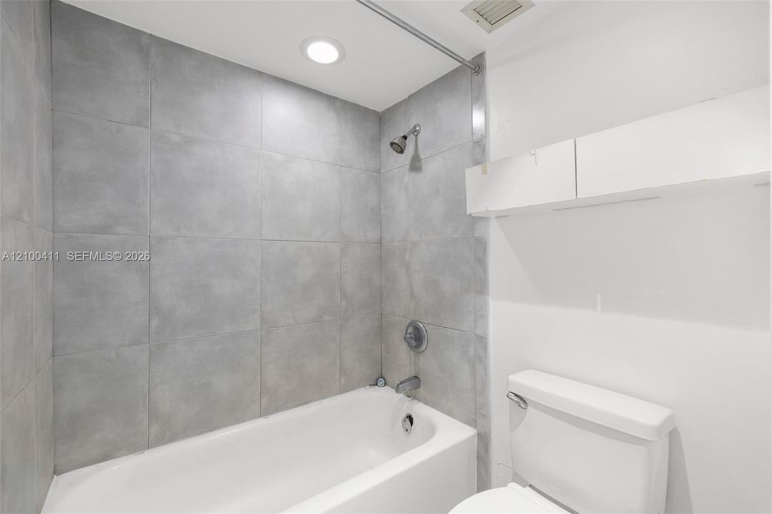Full-height grey tile tub surround, white porcelain bathtub, wall-mounted shower head, recessed ceiling lighting, and built-in white upper cabinetry
