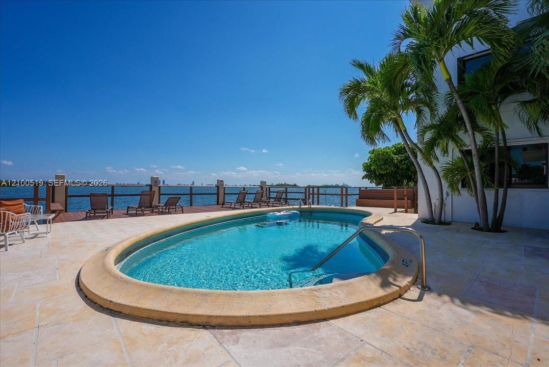 Curved swimming pool with stone coping, expansive stone-paver patio, wooden deck, and waterfront bulkhead