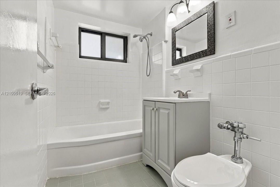 Full bathroom featuring white wall tile, a tub-shower combination with a handheld showerhead, a grey vanity with a white countertop, and a framed mirror with integrated lighting
