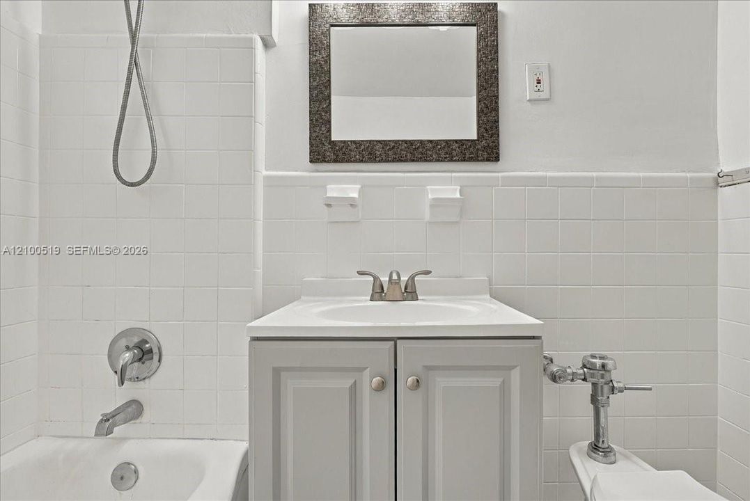 Bathroom featuring white wall tile, grey vanity with integrated sink, chrome dual-handle faucet, wall-mounted mirror with decorative frame, and chrome shower hardware