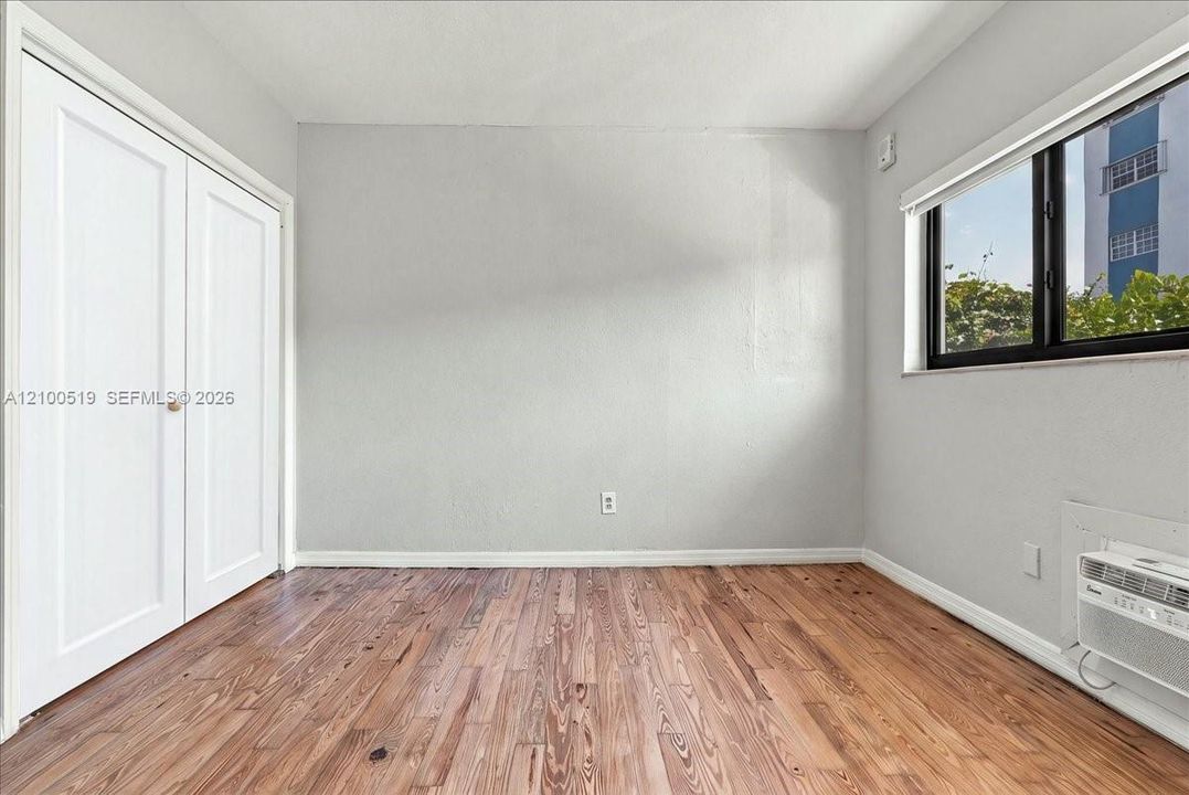 Room featuring wood-finish flooring, white double-door closet, window with black framing, and wall-mounted climate control unit