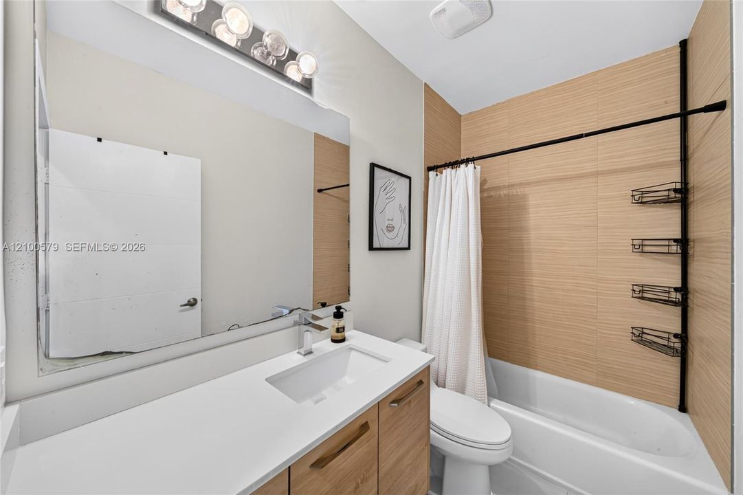 Bathroom featuring a wood-finish vanity with a white rectangular sink, a large wall-mounted mirror with integrated light fixture, and horizontal-tile tub surround