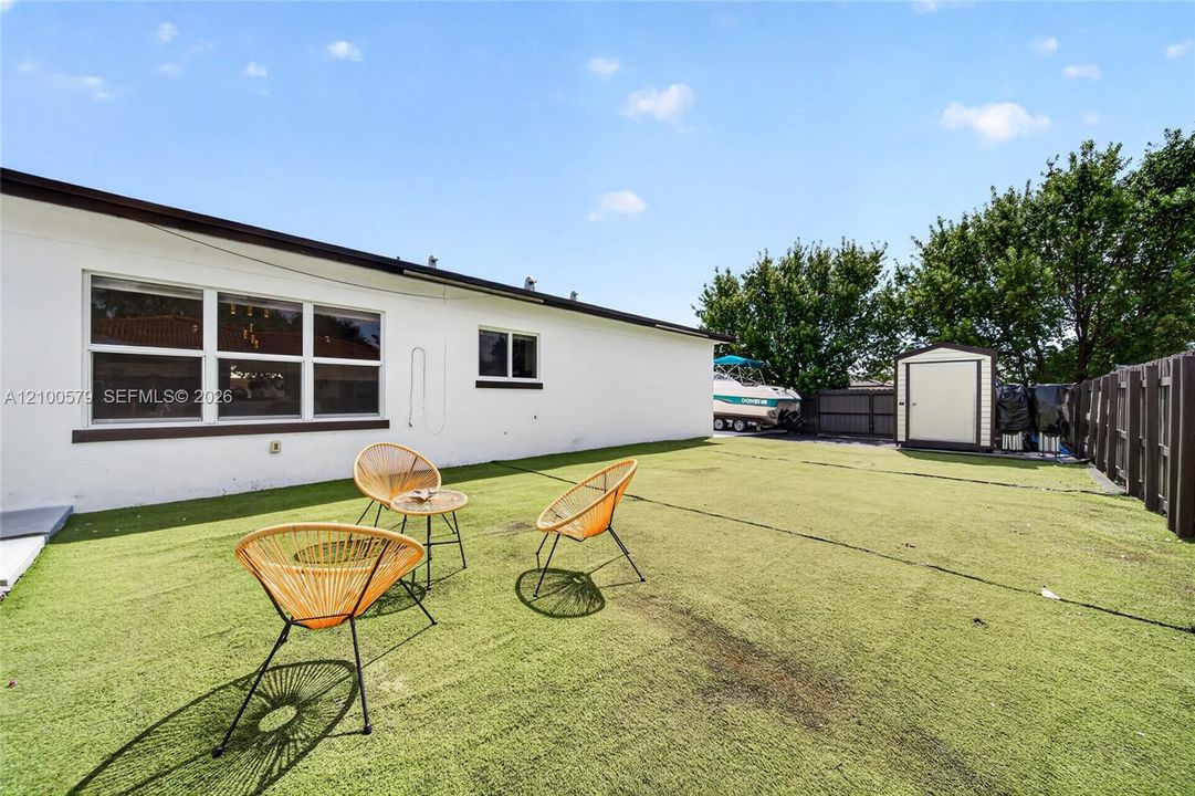 Backyard featuring synthetic turf, white stucco exterior, multipane windows, and a detached storage shed enclosed by wood fencing