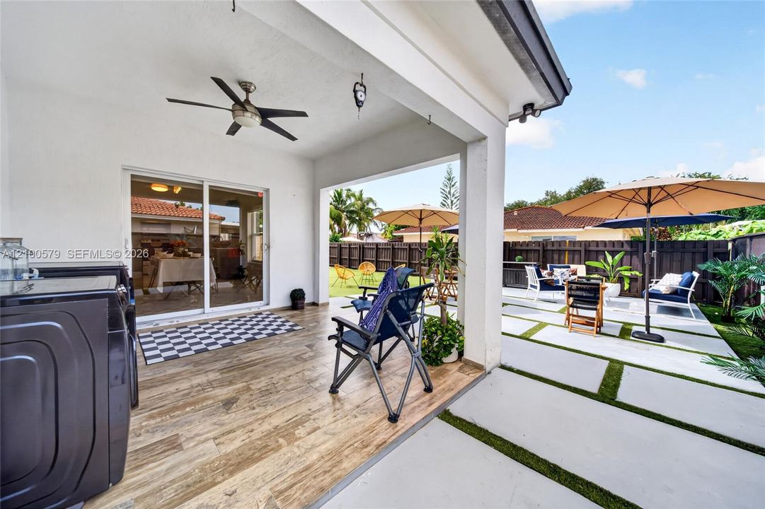 Covered patio featuring wood-finish flooring, a ceiling fan, and sliding glass door access, complemented by an expansive concrete paver patio with turf inlays and a perimeter privacy fence
