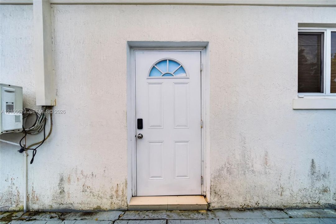 White exterior wall with textured stucco finish, featuring a four-panel entry door with a semi-circular transom window and a rectangular window with a white frame