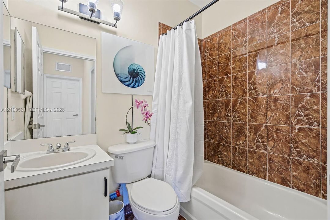 Bathroom featuring a large vanity with an integrated sink, polished chrome dual-handle faucet, expansive wall-mounted mirror, three-light fixture, and shower area with decorative stone-finish tile surround