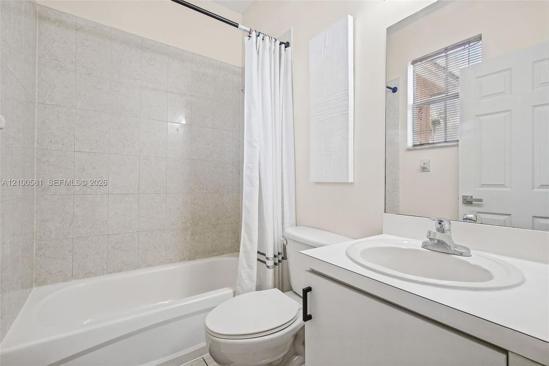 Bathroom featuring a bathtub with full-height tile surround, vanity with an integrated basin, large wall-mounted mirror, and a single window with blinds