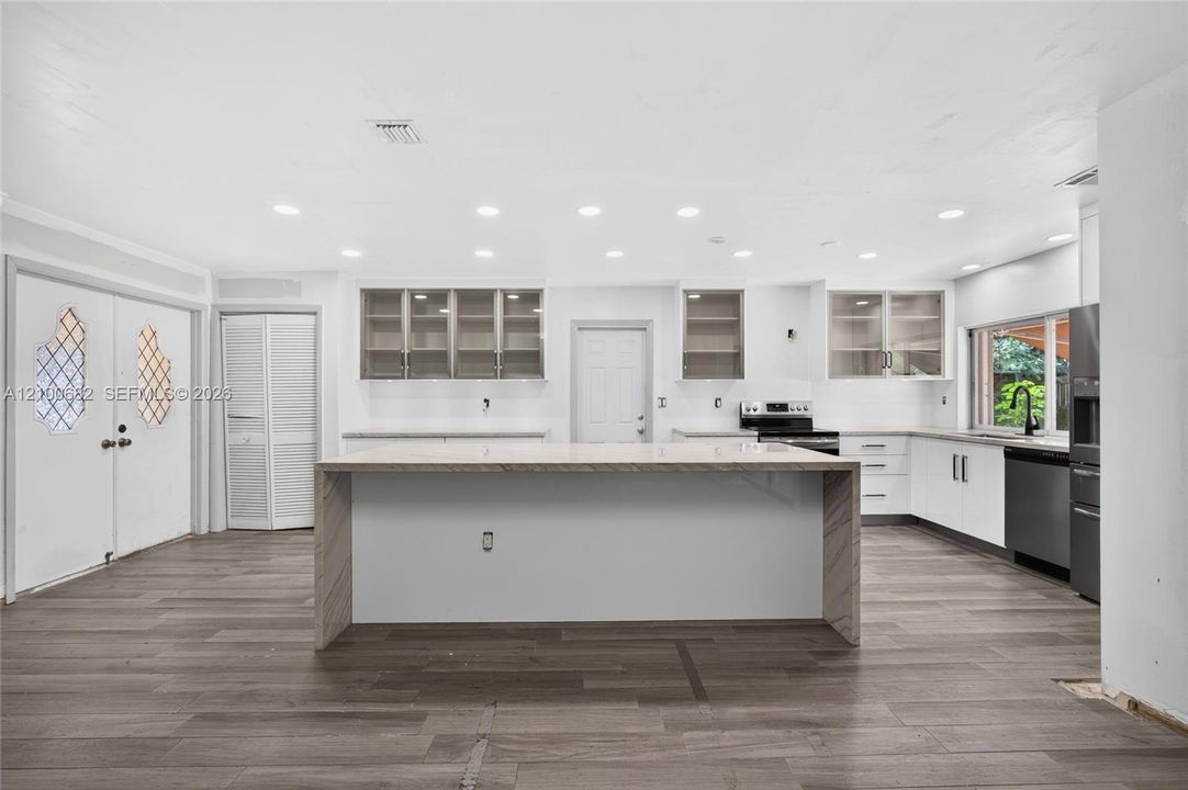 Kitchen featuring a waterfall-edge center island, tiled wood-like flooring, white cabinetry with glass-front uppers, recessed lighting, and stainless steel appliances