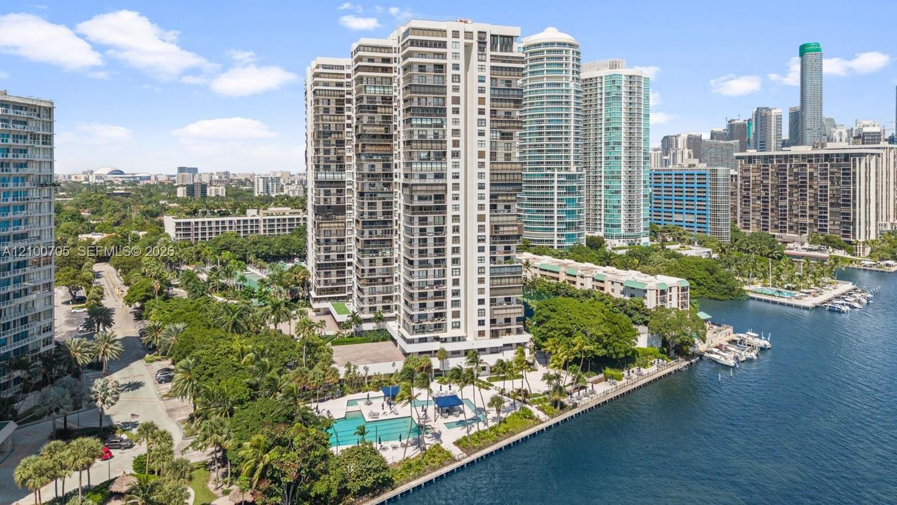 High-rise residential building featuring floor-to-ceiling glass paneling, a ground-level swimming pool, expansive waterfront frontage, and mature landscaping
