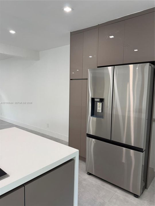 High-gloss cabinetry with floor-to-ceiling storage, stainless steel refrigerator with exterior water dispenser, island featuring a solid-surface countertop, and recessed ceiling lighting