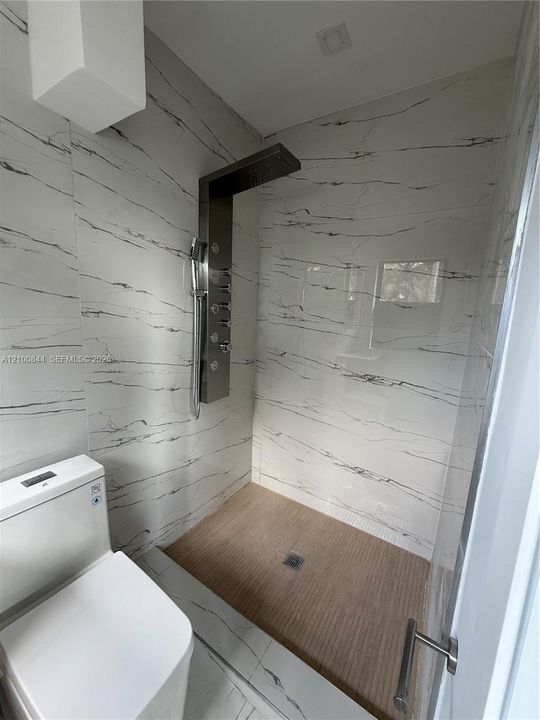 Walk-in shower featuring large-format veined tile surround, brushed metal multi-head shower panel, integrated handheld sprayer, wood-texture floor tiling, and glass door enclosure