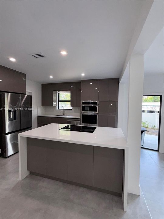 Modern kitchen featuring high-gloss cabinetry, a white waterfall-edge island, a recessed glass cooktop, stainless steel appliances, and large-format tile flooring