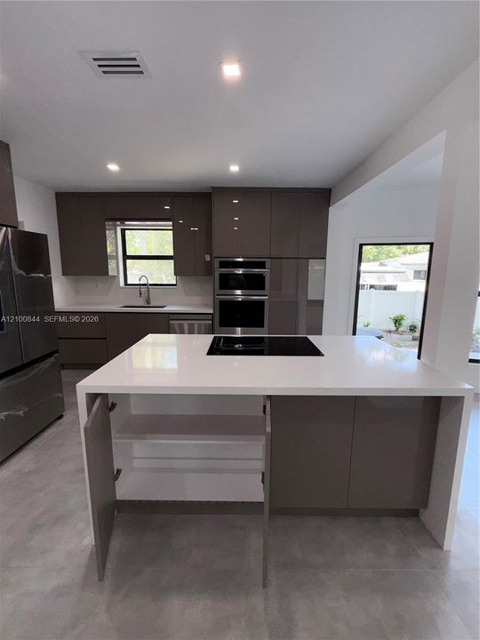 Kitchen featuring a waterfall-edge center island with a cooktop, slab-front cabinetry, stainless steel wall-mounted oven and microwave, recessed lighting, and large-format tile flooring