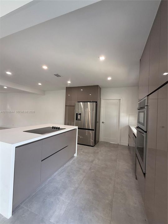Kitchen featuring gloss-finish cabinetry, an island with a waterfall-edge countertop and integrated cooktop, large-format tile flooring, and stainless steel built-in appliances including a wall oven and refrigerator