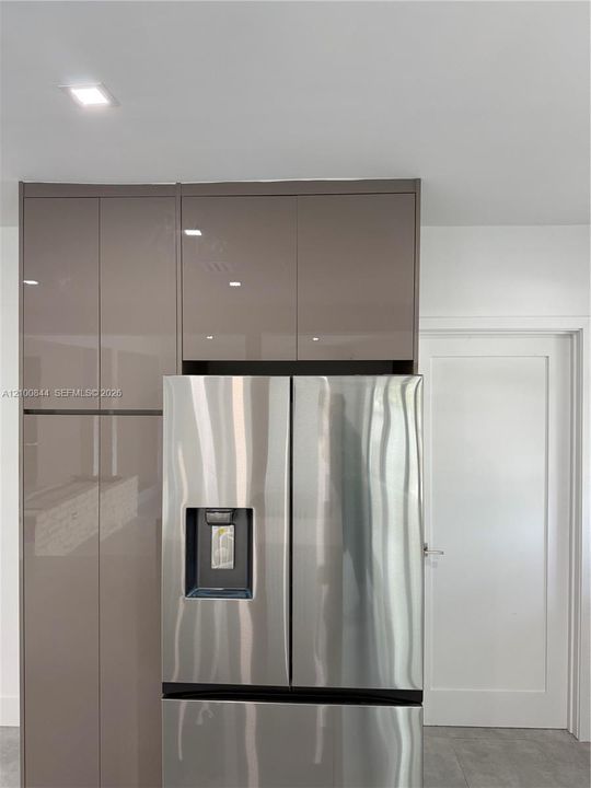 High-gloss taupe cabinetry featuring a built-in refrigerator enclosure, stainless steel refrigerator with external water dispenser, recessed ceiling lighting, and a single-panel white interior door