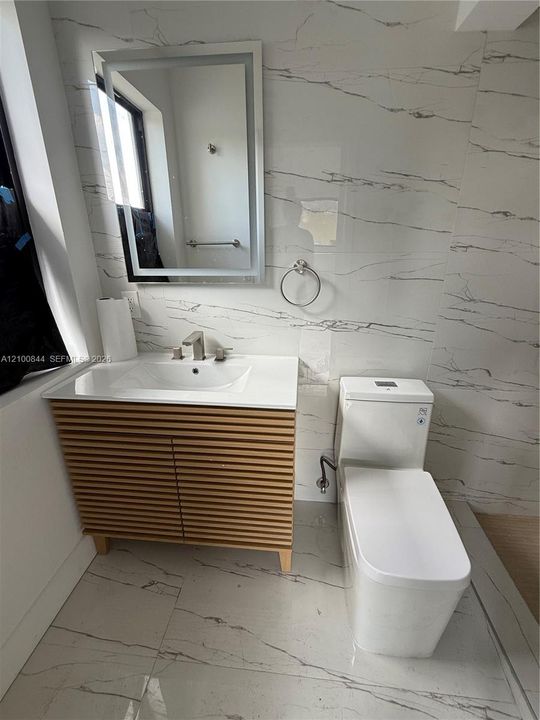 Slatted wood-finish vanity with a white countertop and integrated sink, illuminated LED mirror, floor-to-ceiling veined stone-finish tile, and a rectangular dual-flush toilet